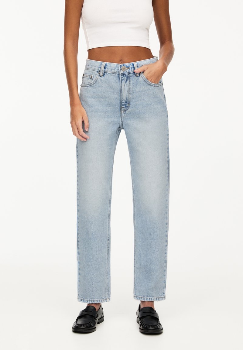 Femme portant un jean droit taille haute bleu clair, un top court blanc et des mocassins noirs, debout avec une main dans la poche.