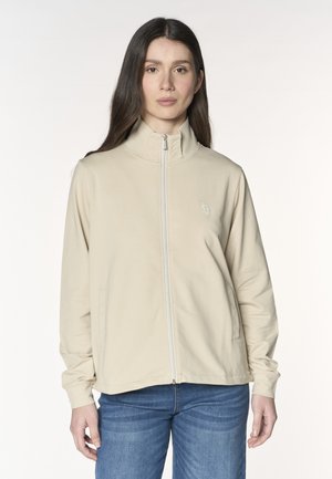 Donna che indossa una giacca beige con zip e collo alto e jeans blu, in piedi su uno sfondo bianco uniforme.
