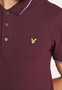Burgundy polotröja med tvåknapps-polo, grå kant och en liten gul logotyp broderad på vänster bröst. Texturerad tygdetalj.