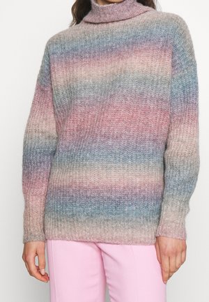 Personne portant un pull à col roulé côtelé aux couleurs pastel multicolores et un pantalon rose clair, debout devant un fond blanc uni.