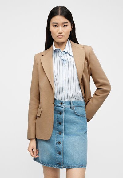 Γυναίκα που φοράει καμηλό σακάκι, λευκό πουκάμισο με ρίγες και μπλε τζιν φούστα με κουμπιά στο μπροστινό μέρος, στέκεται με το ένα χέρι στη μέση της.