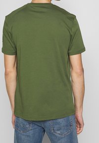 Man som bär en enkel olivgrön kortärmad t-shirt i kombination med blå denimjeans, vänd bort, som visar bakviewen av outfiten.