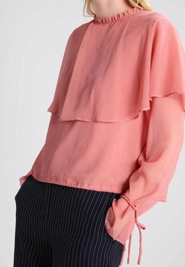 Femme portant un chemisier corail à volants avec col à volant et poignets à nouer, associé à un pantalon sombre à fines rayures.