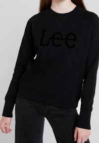 Sudadera negra de manga larga con el logo negro de "Lee" usada con jeans negros por una persona contra un fondo claro y plano.