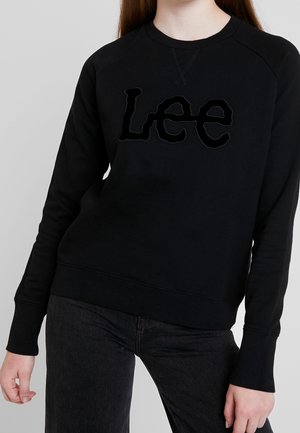 Felpa nera a maniche lunghe con logo "Lee" nero indossata con jeans neri da una persona su uno sfondo chiaro e semplice.