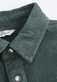 Zielony kołnierzyk koszuli z korduroju z pionowymi żłobieniami, srebrne guziki i widoczna metka. Teksturowana tkanina nadaje miękki wygląd.