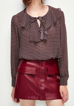 Kvinde iført en mønstret bluse med flæsekrave og en knapforan vinrød lædermininederdel med klaplommer.