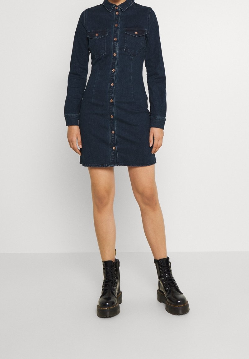Abito camicia di jeans scuro con bottoni frontali, due tasche sul petto, maniche lunghe e silhouette aderente, abbinato a stivali neri con lacci.