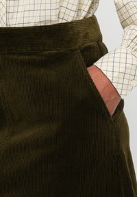 Zöld kordbársony szoknya, széles derékrésszel és oldalzsebekkel, függőleges bordázott textúrával. Kockás inggel viselve.