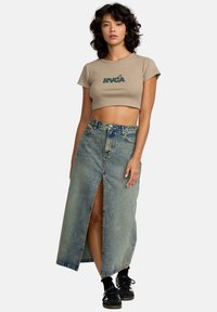 Kuttet beige t-skjorte med grønt logo, parret med et langt denimskjørt med frontslitt, blå vask og kontrasterende stikking. Svarte joggesko.