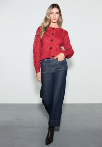 Red cable-knit cardigan with black buttons, paired with wide-leg dark denim pants. Black heeled ankle boots complete the outfit.
