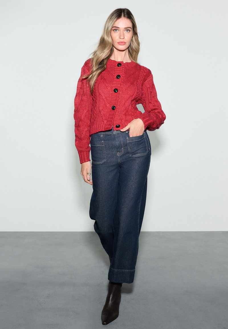 Red cable-knit cardigan with black buttons, paired with wide-leg dark denim pants. Black heeled ankle boots complete the outfit.