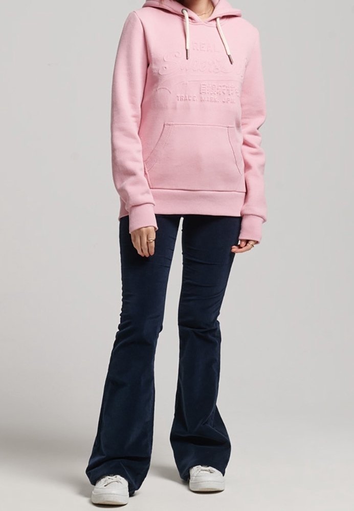 Sudadera rosa claro con un bolsillo frontal y capucha con cordón, combinada con pantalones acampanados oscuros navy y zapatillas blancas. Tejido suave y texturizado.