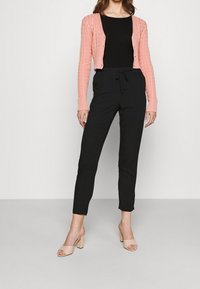 Kvinna iklädd en rosa kortärmad kabelstickad cardigan över en svart topp, svarta dragsko-byxor och beiga öppna blockklackar står på ett ljust golv.