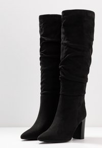 Bottes hautes jusqu'au genou en daim noir avec texture froncée, bout pointu et talon carré. Extérieur lisse avec une finition douce.