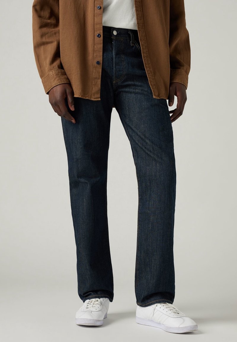 Personne portant un jean droit bleu foncé, des baskets blanches, une chemise blanche et une chemise marron boutonnée ouverte sur un fond uni.