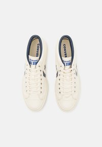 Bež platnene tenisice s mornarskim naglaskom, s okruglim vrhom, ravnim vezicama i zvjezdastim logotipom sa strane. Tekst glasi "Converse All Star."