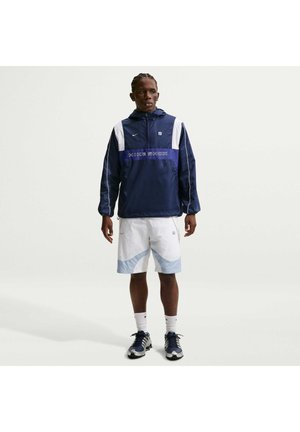 Mand stående iført marineblå og hvide Nike Shox vindjakke, hvide og lyseblå shorts, hvide sokker og marineblå sneakers på ensfarvet baggrund.