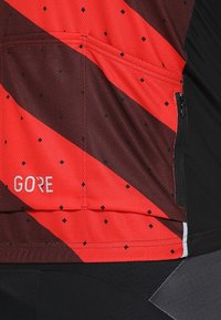Jersey de ciclismo rojo y negro con rayas diagonales, tejido texturizado, bolsillo con cremallera y pequeños patrones de diamantes. Logo de GORE exhibido.