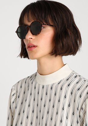 Jonge vrouw met kort donker haar die ronde zonnebrillen van schildpadmotief draagt en een witte blouse met hoge hals en zwarte verticale patronen.