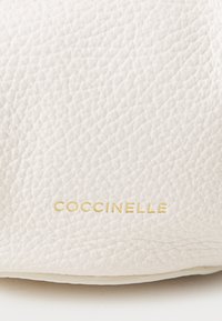 Pelle bianca testurizzata con logo "Coccinelle" in oro in rilievo. Dettaglio ravvicinato che mette in evidenza il materiale e il marchio.