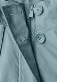 Hellblaue Stoffjacke mit sichtbarem Reißverschluss, großem Knopf und genähten Akzenten. Glatte Textur mit sauberem Finish.