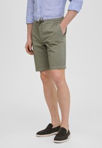 Shorts de algodón verde oliva con cintura elástica y cordón, bolsillos laterales y corte recto, combinados con zapatos negros de tipo slip-on.