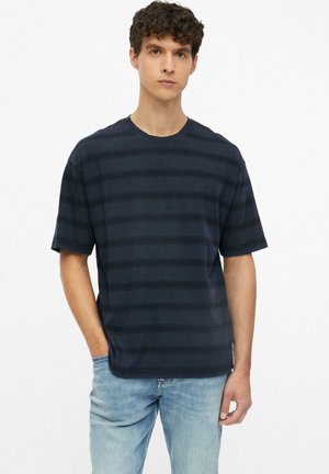 Junger Mann mit lockigem Haar trägt ein dunkelmarineblau gestreiftes kurzärmeliges T-Shirt und hellblaue Jeans, steht mit einer Hand in der Tasche.