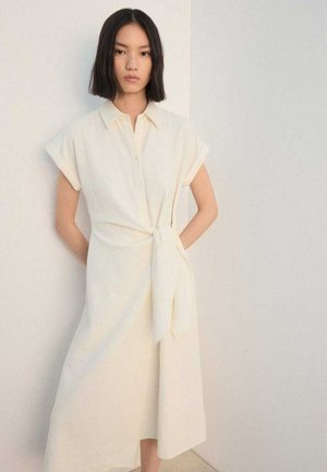 Roomkleurige overhemdjurk met korte opgerolde mouwen, knopen aan de voorkant en een strikdetail in de taille. Gemaakt van lichtgewicht stof met een gestructureerde afwerking.