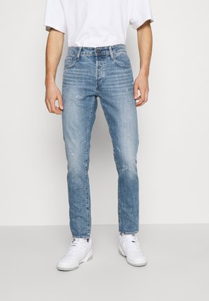 Mann trägt hellblaue Slim-Fit-Jeans, weiße Turnschuhe und ein schlichtes weißes kurzärmeliges T-Shirt, steht vor einem neutralen Hintergrund.