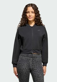 Crna skraćena dukserica na cipz za dugim rukavima; sadrži suptilan logo na prsima. U kombinaciji s crnim tajicama s leopard uzorkom.