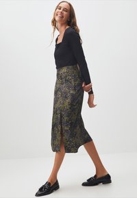 Zwarte lange mouwen top gecombineerd met een middellange rok in groen en zwart luipaardprint, met een split aan de voorkant en elegante loafers.