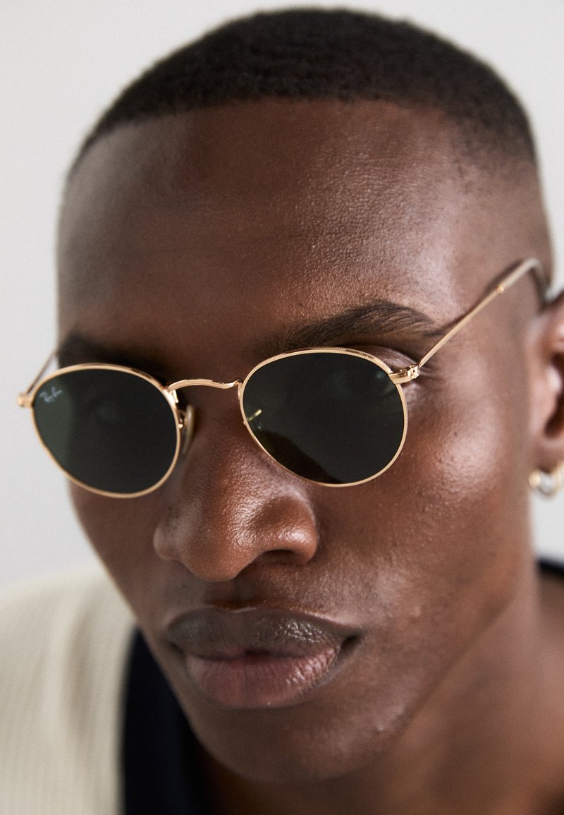 Ochelari de soare rotunzi cu ramă aurie și lentile închise. Brațe subțiri din metal și branding discret lângă lentile. Textură netedă și design minimalist.