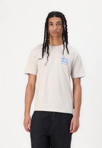 Šviesiai rudas medvilnės marškinėliai trumpomis rankovėmis, su mėlynos bangos dizainu kairėje krūtinės pusėje. Dėvimi su juodomis kelnėmis. Lygi tekstūra.