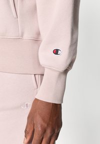 Sweatshirt à capuche rose clair avec des poignets côtelés, présentant un logo brodé en noir, rouge et blanc sur la manche gauche. Tissu doux et texturé.