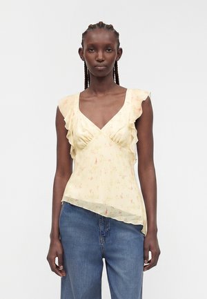Femme aux cheveux tressés portant un haut sans manches à volants jaune clair à motif floral et un jean bleu, debout devant un fond uni.
