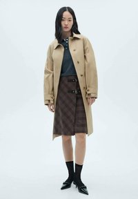 Trench coat bej cu nasturi mari, asortat cu un polo gri închis și o fustă cu carouri maro. Sosetele negre până la glezne și balerinii ascuțiți completează ținuta.