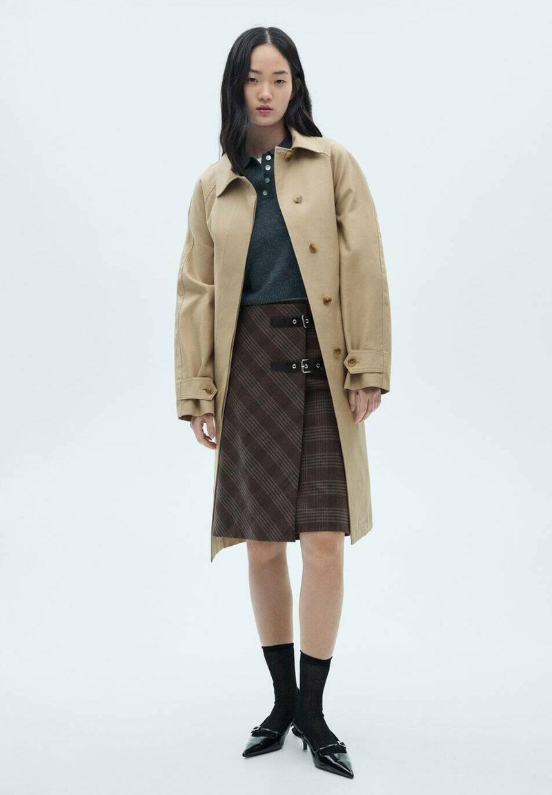 Trench coat bej cu nasturi mari, asortat cu un polo gri închis și o fustă cu carouri maro. Sosetele negre până la glezne și balerinii ascuțiți completează ținuta.