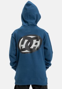 Mikina s kapucňou v tmavomodrom bavlnenom materiáli, na zadnej strane s veľkým čierno-bielym logom. Mäkky materiál, voľný strih a dlhé rukávy.