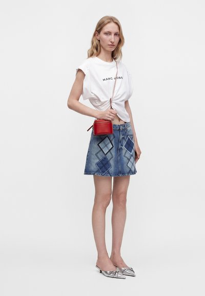 Γυναίκα που φοράει λευκό t-shirt Marc Jacobs, μπλε denim μίνι φούστα με διακοσμητικά διαμάντια, ασημένιες μυτερές γόβες και κρατά μια μικρή κόκκινη τσάντα χιαστί.