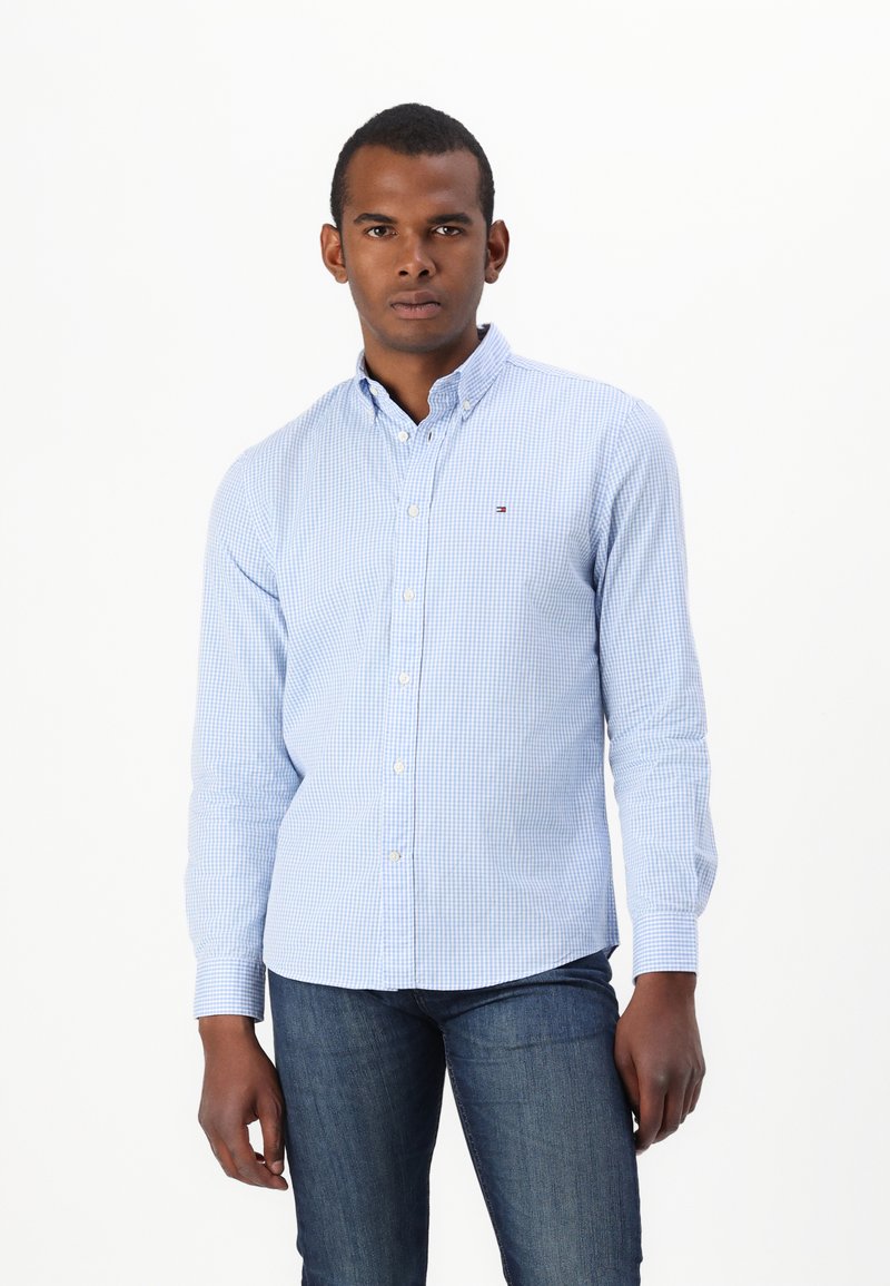 Camicia button-up a quadri blu chiaro con colletto a punta, maniche lunghe e un piccolo logo sul petto, abbinata a jeans scuri.
