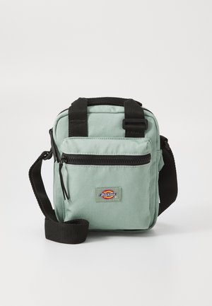 Világoszöld anyagú, keresztpántos táska fekete pántokkal, elülső cipzáras zsebbel és logóval ellátott folttal. Téglalap alakú, kompakt dizájn, és tartós textúra.