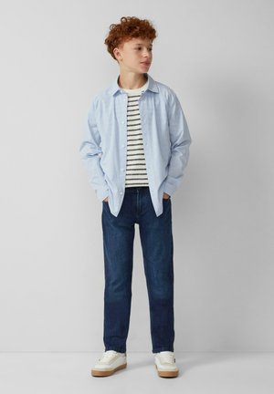 Lichtblauw gestreept overhemd over een zwart-wit gestreepte t-shirt, gecombineerd met donkere jeans en witte sneakers met een tan kleurige zool.