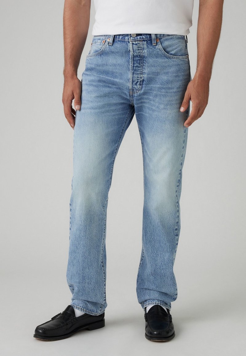 Homme portant un jean bleu clair délavé, une chemise blanche, des chaussettes blanches et des mocassins noirs, debout devant un fond gris uni.