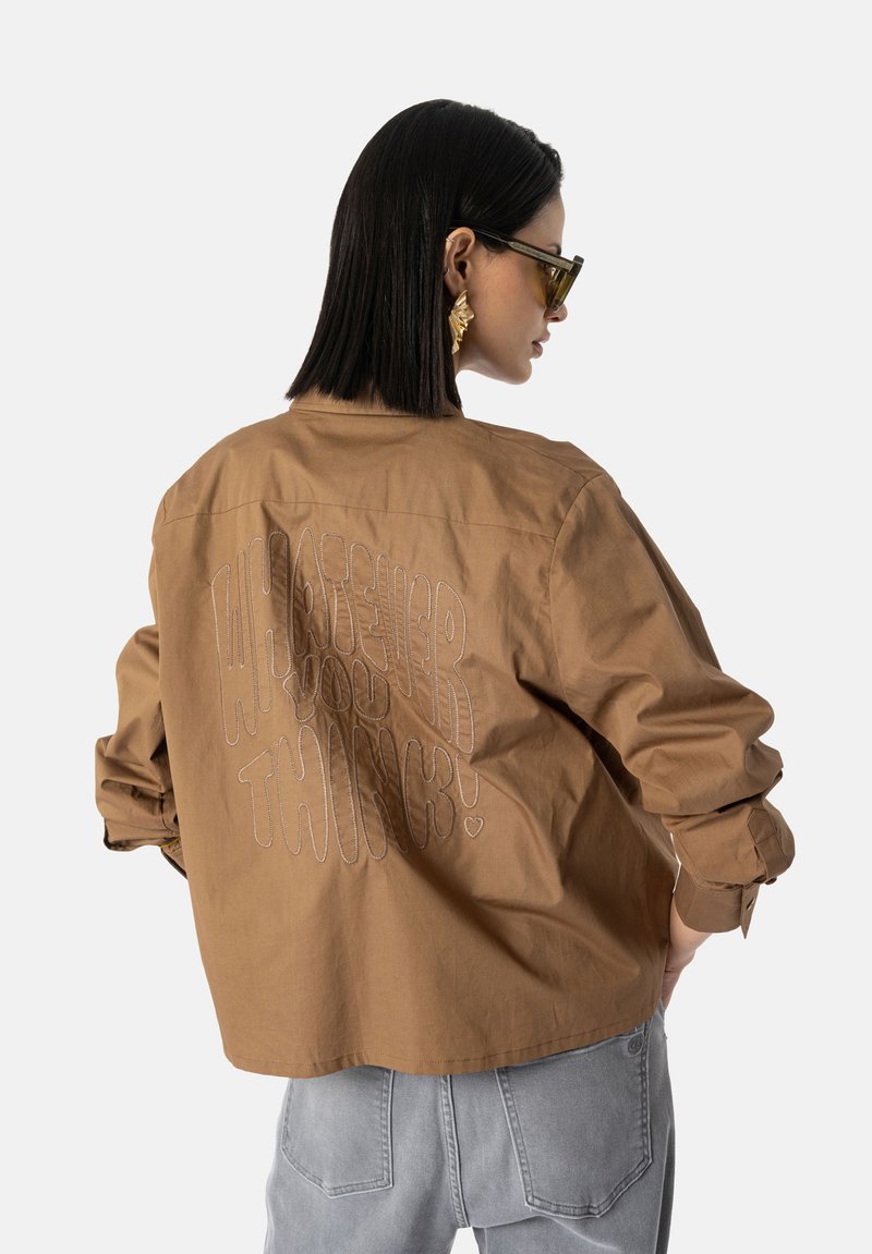 Bruin katoenen jack met een relaxte pasvorm, voorzien van een grote witte geborduurde tekst "WAT JE OOK DENKT!" op de rug. Lange mouwen, knoopmanchetten.