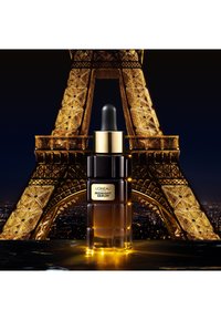 Sérum de nuit L'Oréal dans un flacon en verre sombre avec un compte-gouttes, présentant un design texturé et des accents dorés, exposé devant la Tour Eiffel.