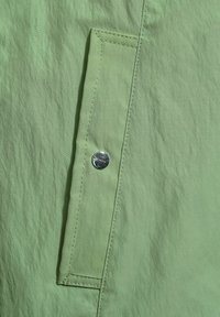 Ljusgrön tyg med en texturerad yta, en sidficka med synlig sömnad och en detalj med silvertryckknapp.