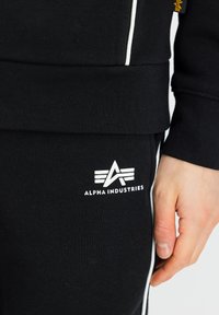 Zwarte sweatshirt en broek met witte zijstrepen. Bevat een wit logo met de tekst "ALPHA INDUSTRIES" op de broek. De stof lijkt soepel.