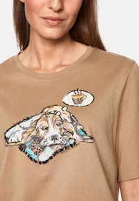 Braunes Baumwoll-T-Shirt mit einem Grafikmotiv eines Beagle auf einer Decke und einer Sprechblase mit einer gemusterten Kaffeetasse. Kurze Ärmel.