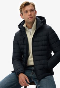 Chaqueta acolchada negra con capucha, que presenta un cierre frontal y puños elásticos, llevada sobre un suéter de cuello alto de color claro y jeans.